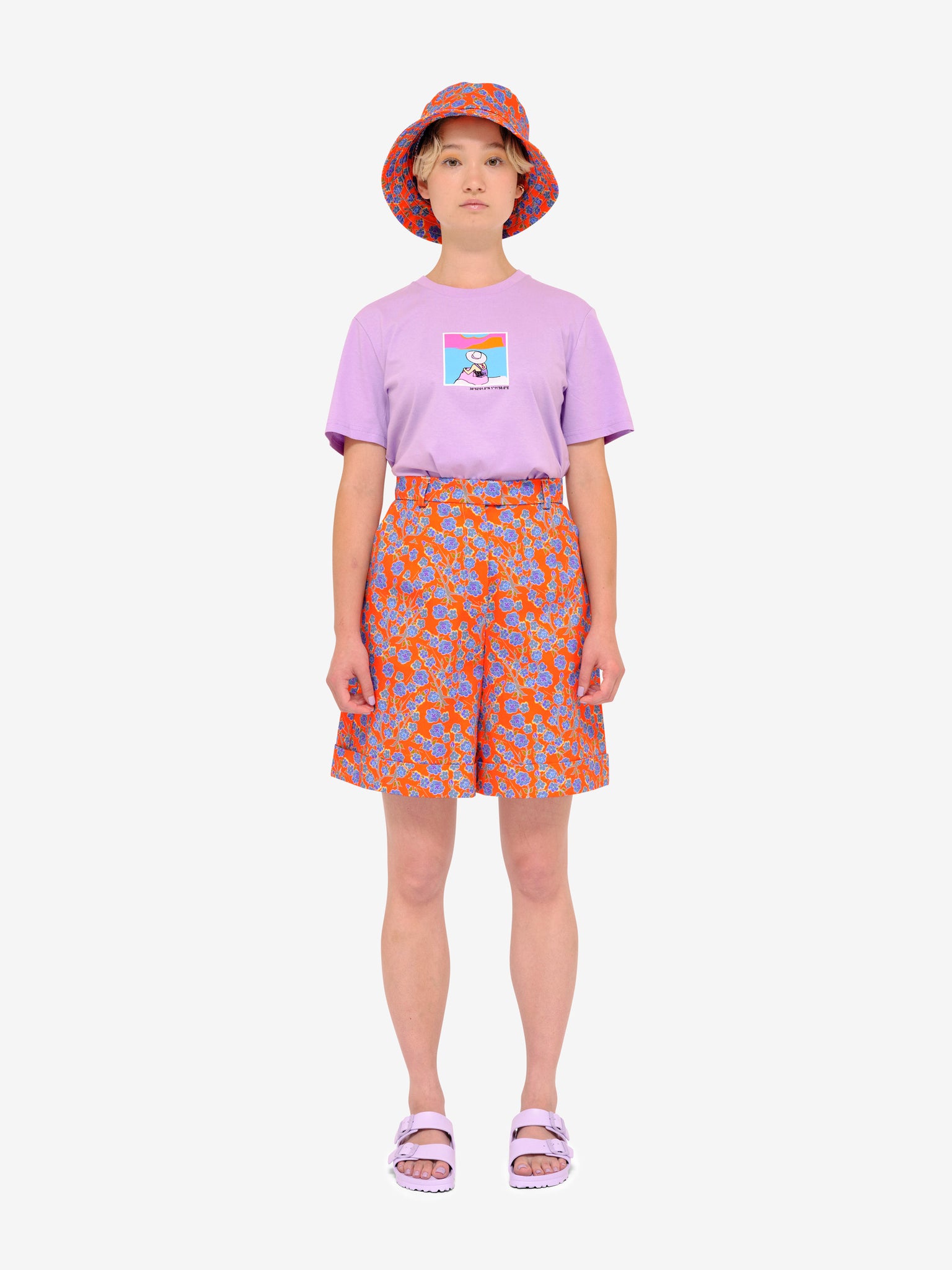 Model wearing Crimson Rose Es Vedrà lilac T-shirt with orange floral print shorts and bucket hat and lilac sandals. Photography Rowan Corr.